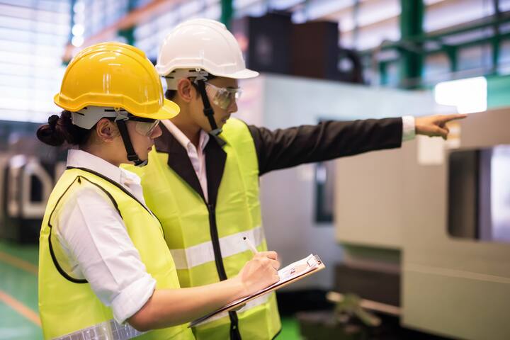 Waist up female factory worker write implement audit result of industrial machine while manager point finger to machinery. Manufacturing industry to produce car spare parts. Quality assurance (QA).