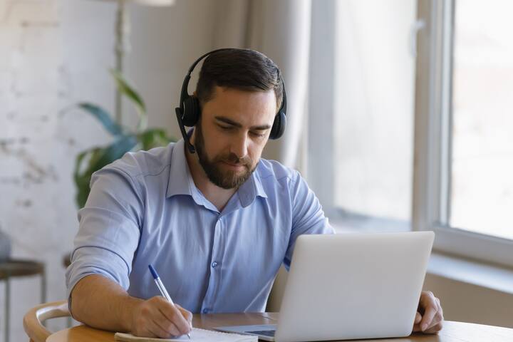 Focused young man in headset with microphone listening educational lecture on computer, studying on online courses at home, writing notes improving professional knowledge, preparing for exams.