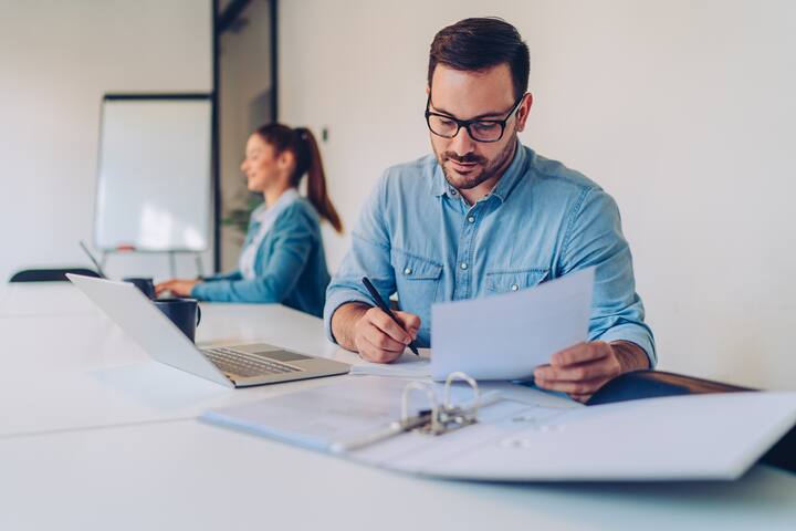 Young businessman wearing denim shirt and eyeglasses doing paperwork while sitting at the desk in the big white office with coworkers