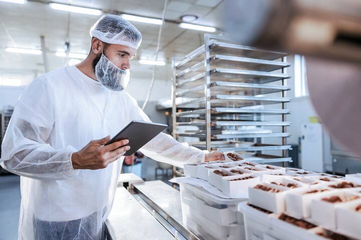 A food inspector in a sterile white uniform is holding the tablet and looking at collected cookies. Food check is important if we want quality and healthy food.