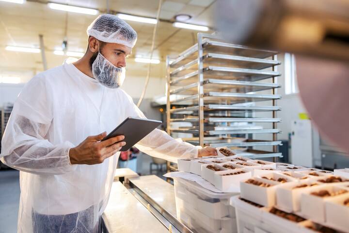 A food inspector in a sterile white uniform is holding the tablet and looking at collected cookies. Food check is important if we want quality and healthy food.