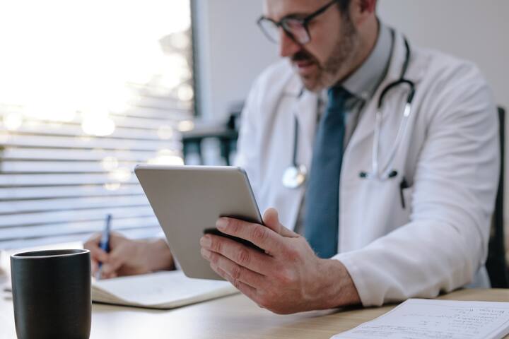 Mature male doctor holding a digital tablet making notes in his diary while sitting in his office. Doctor sitting at his desk with tablet computer and writing prescription for his patient. Focus on digital tablet in hand of doctor.