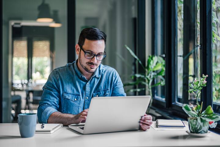 Serious businessman using laptop while working at table. Male entrepreneur wearing denim shirt. He is doing freelance work at home office.