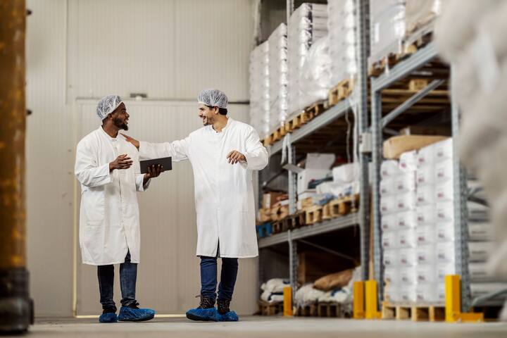 Full length shot of food plant warehouse workers with tablet walking towards camera and talking. Team of two diverse food factory workers collaborating