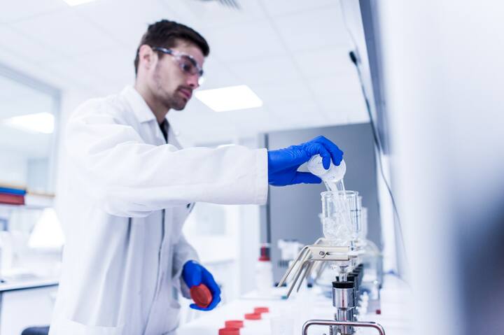 Man wearing a lab coat and safety goggles conducts a chemical test by pouring a liquid into a glass filtration setup, wearing blue gloves in a modern laboratory.