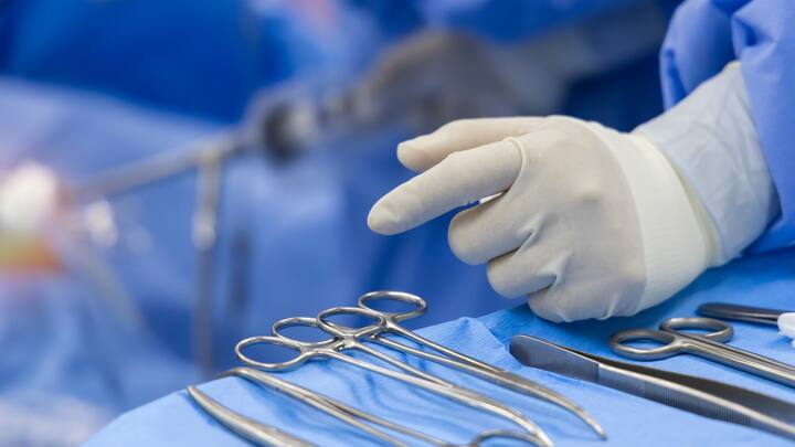 A gloved hand selects a surgical instrument from a sterile set in an operating room. Normec.