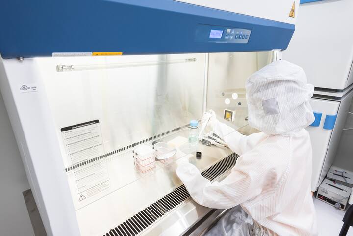 A scientist in sterile coverall gown pipetting medium or reagents for cell culture experiment in biological safety cabinet. Doing biological research in clean environmental. Cleanroom facility