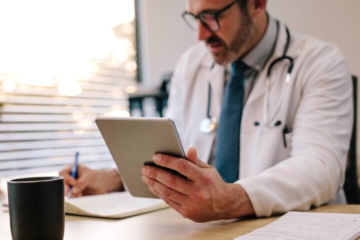 Mature male doctor holding a digital tablet making notes in his diary while sitting in his office. Doctor sitting at his desk with tablet computer and writing prescription for his patient. Focus on digital tablet in hand of doctor.