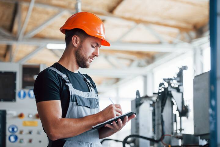 With notepad in hands. Industrial worker indoors in factory. Young technician with orange hard hat.