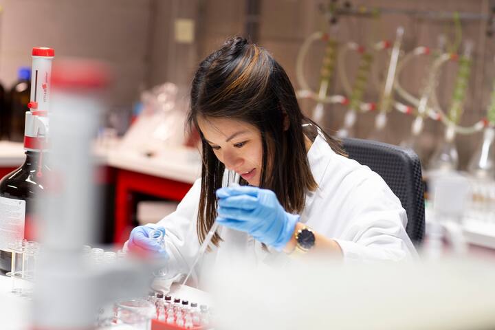 Female lab technician in a white coat and blue gloves conducting precise pipetting test in a pharmaceutical laboratory at Synergy Health Utrecht.