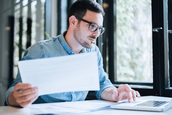 Serious pensive thoughtful focused young casual business accountant bookkeeper in office looking at and working with laptop and income tax return papers and documents